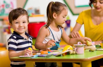 Two children happily playing at a table, with a teacher in the background 