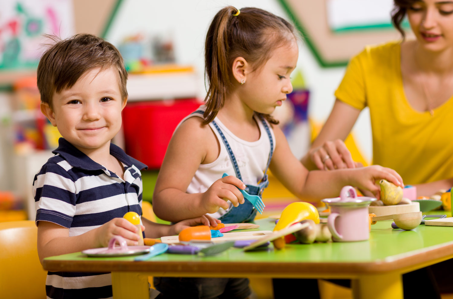 Image of Two children playing at a table, with a teacher in the background